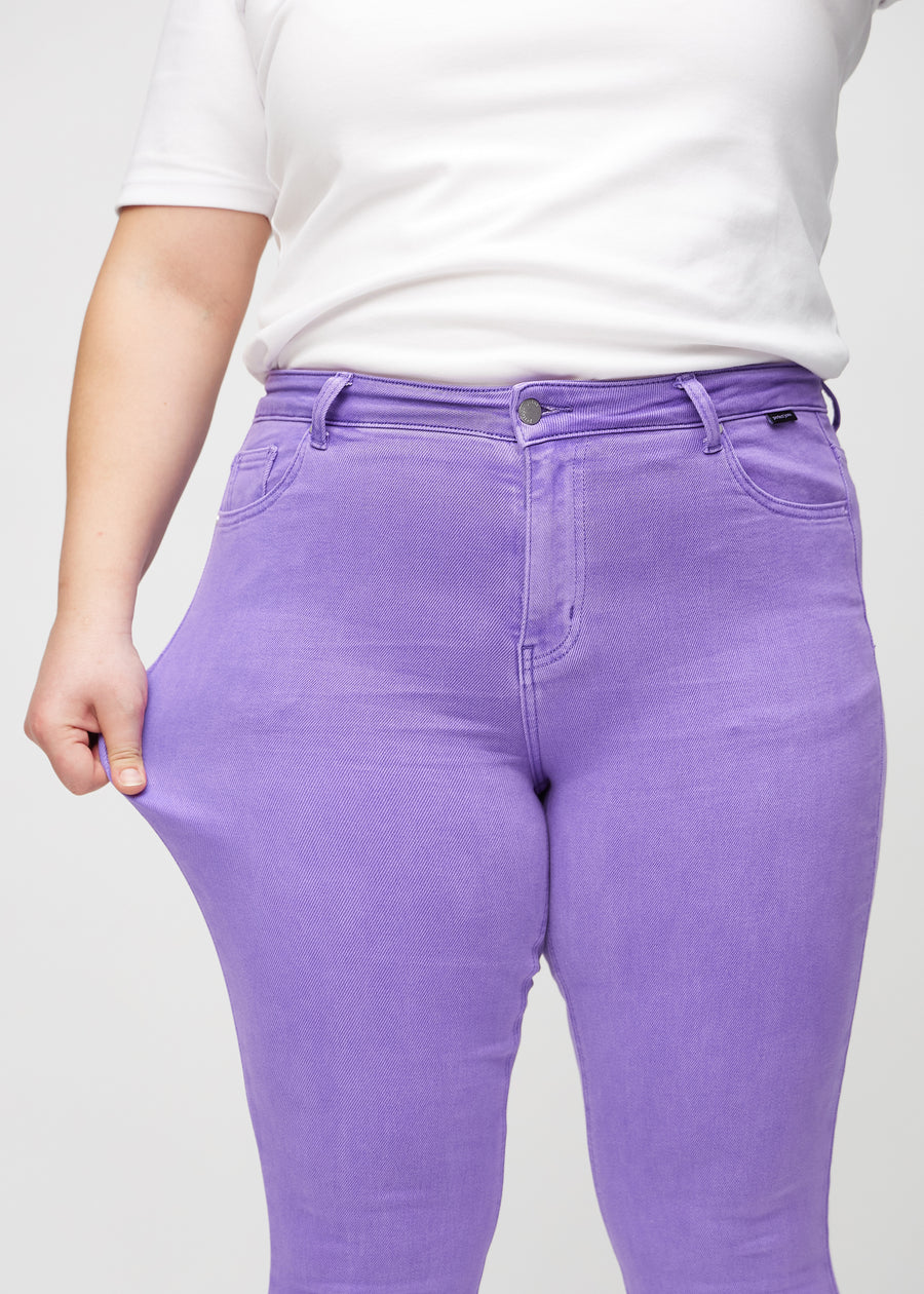 Skinny-jeans til kvinner i lilla, fargen Lavenders. En plus size-modell strekker jeansstoffet ved låret for å vise stretchen.