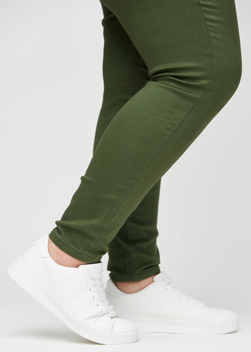 Skinny-jeans til kvinner i mørkegrønn, fargen Renewals. Nærbilde ved anklene på en plus size-modell, som viser de tettsittende benåpningene.