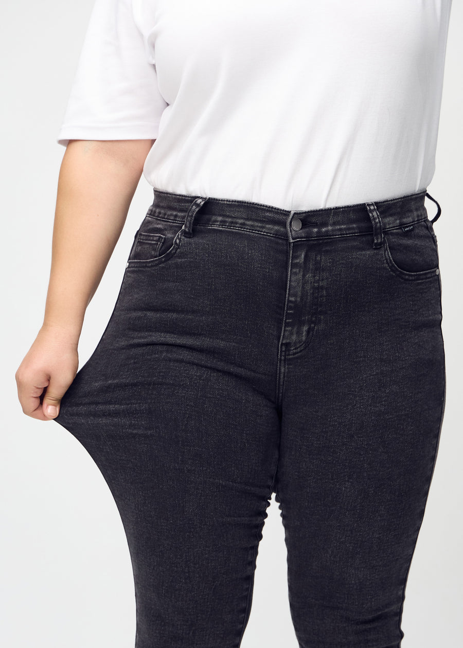 Skinny-jeans til kvinner i mørkegrå, fargen Shadows. En plus size-modell strekker jeansstoffet ved låret for å vise stretchen.