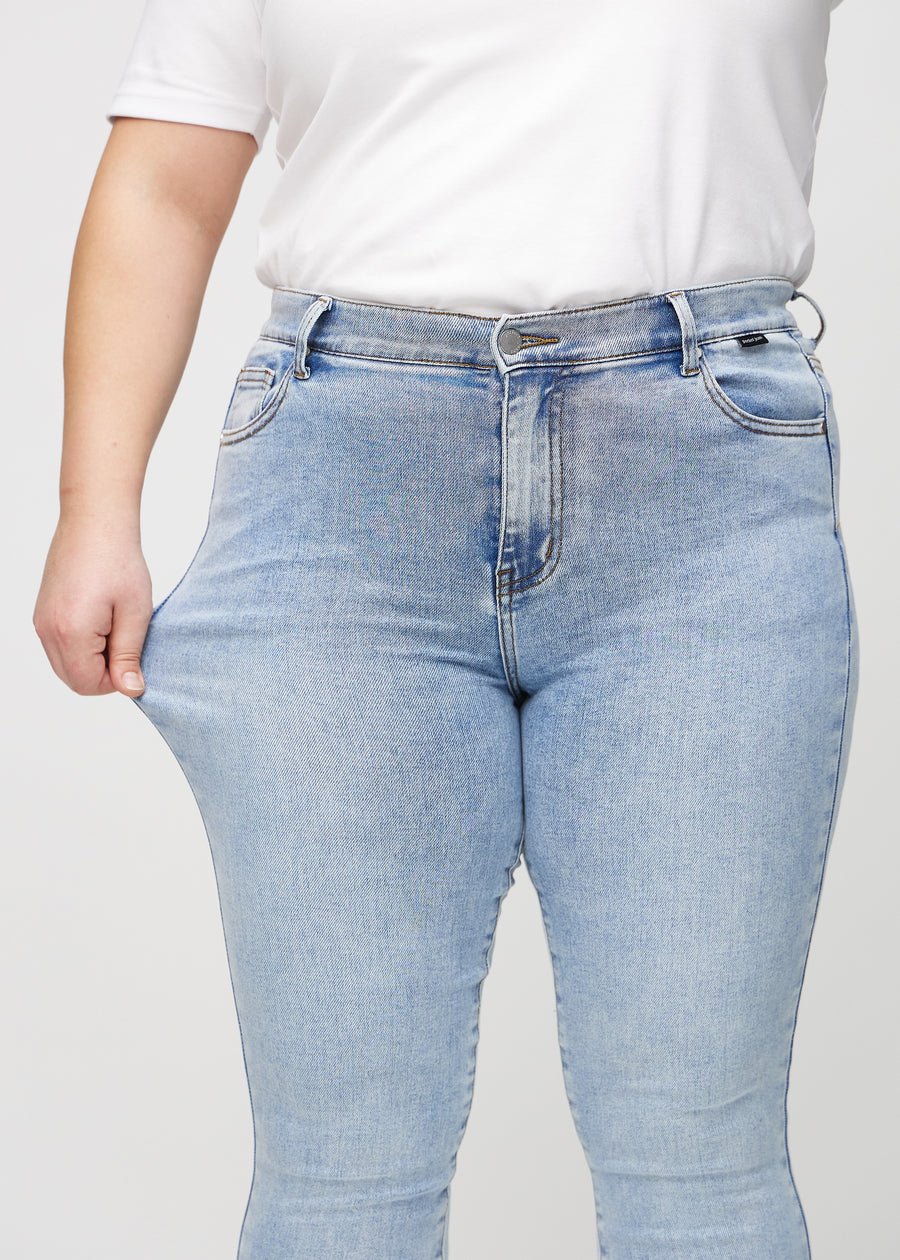 Skinny-jeans til kvinner i lyseblå, fargen Waves. En plus size-modell strekker jeansstoffet ved låret for å vise stretchen.