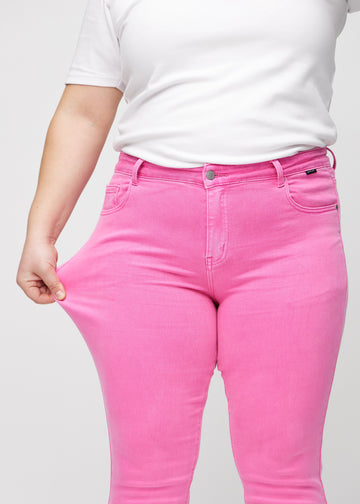 Slim-jeans til kvinner i lyserosa, fargen Flamingos. En plus size-modell strekker jeansstoffet ved låret for å vise stretchen.