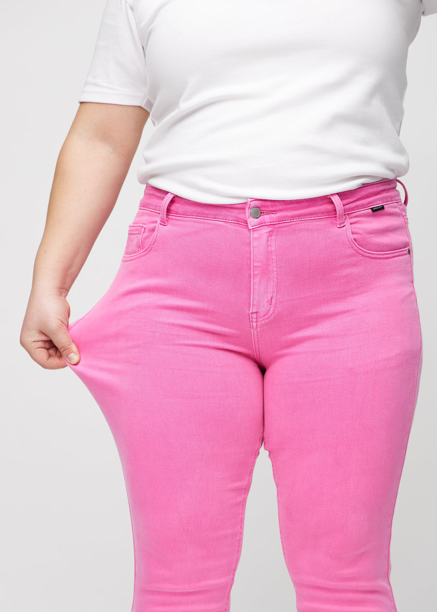 Slim-jeans til kvinner i lyserosa, fargen Flamingos. En plus size-modell strekker jeansstoffet ved låret for å vise stretchen.