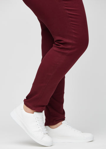 Slim-jeans til kvinner i mørkerød, fargen Santas. Nærbilde ved anklene på en plus size-modell, som viser passformen nederst på bena.