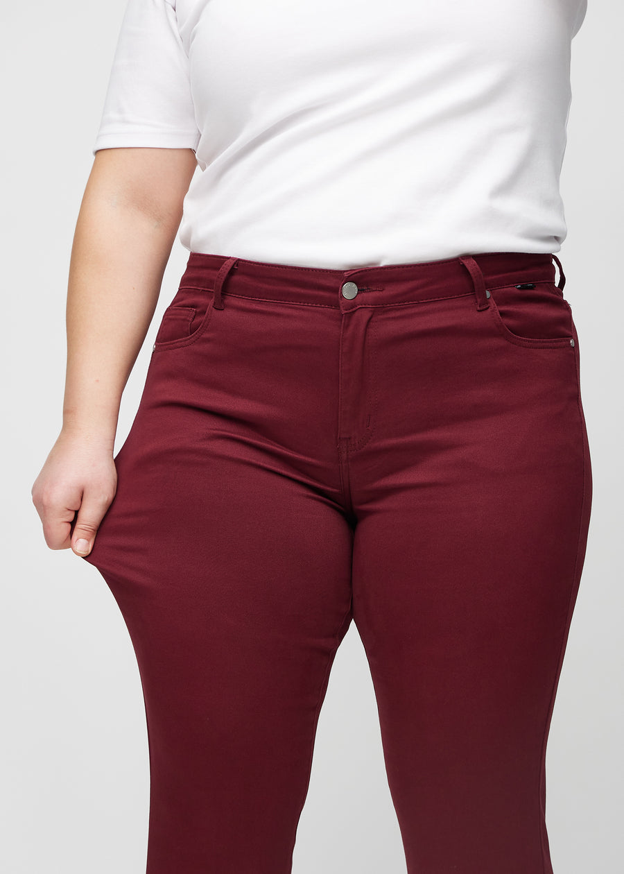 Slim-jeans til kvinner i mørkerød, fargen Santas. En plus size-modell strekker jeansstoffet ved låret for å vise stretchen.