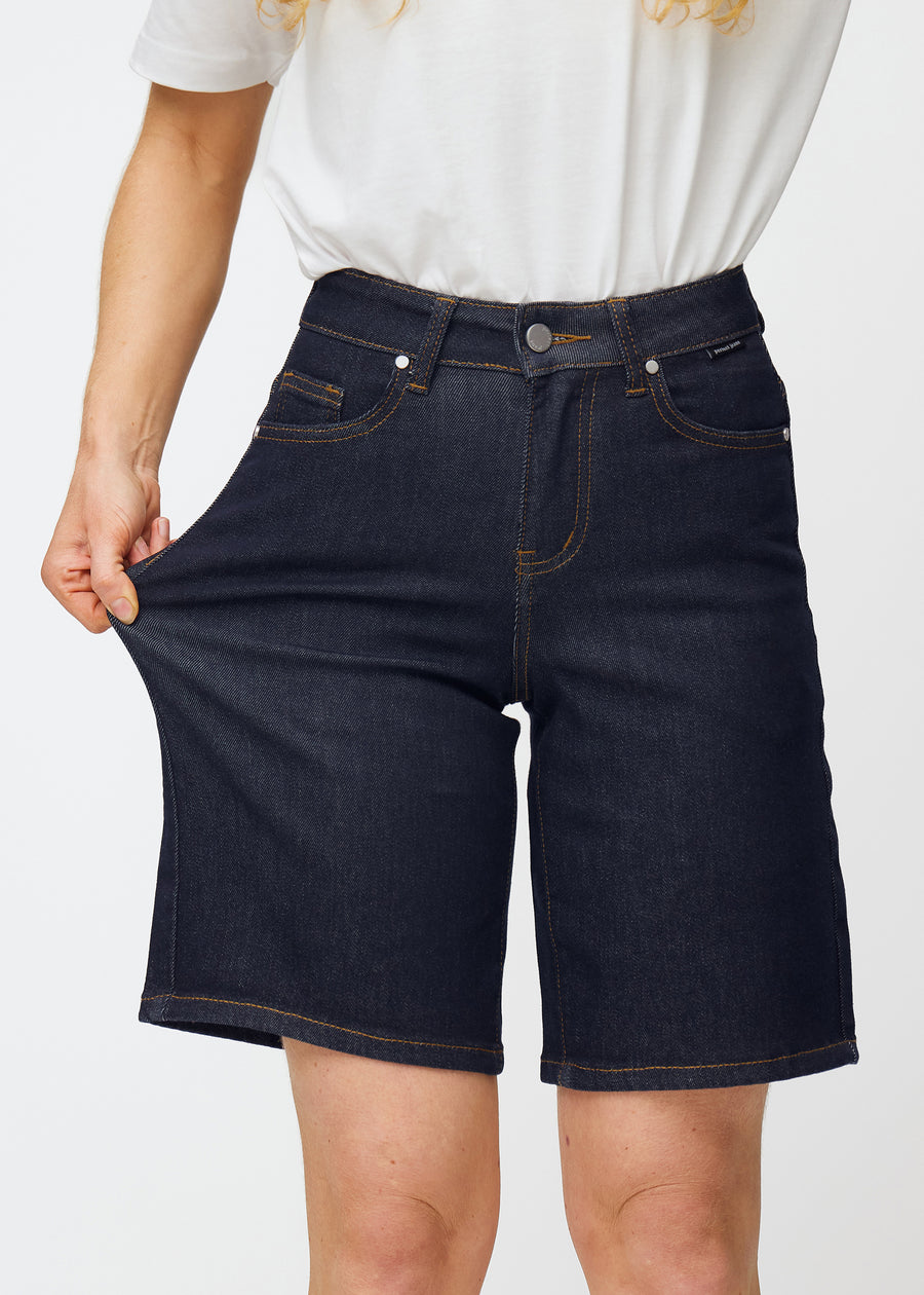 Modell strekker dyp mørkeblå denimshorts til dame i fargen Midnights ved låret for å vise materialets stretch.