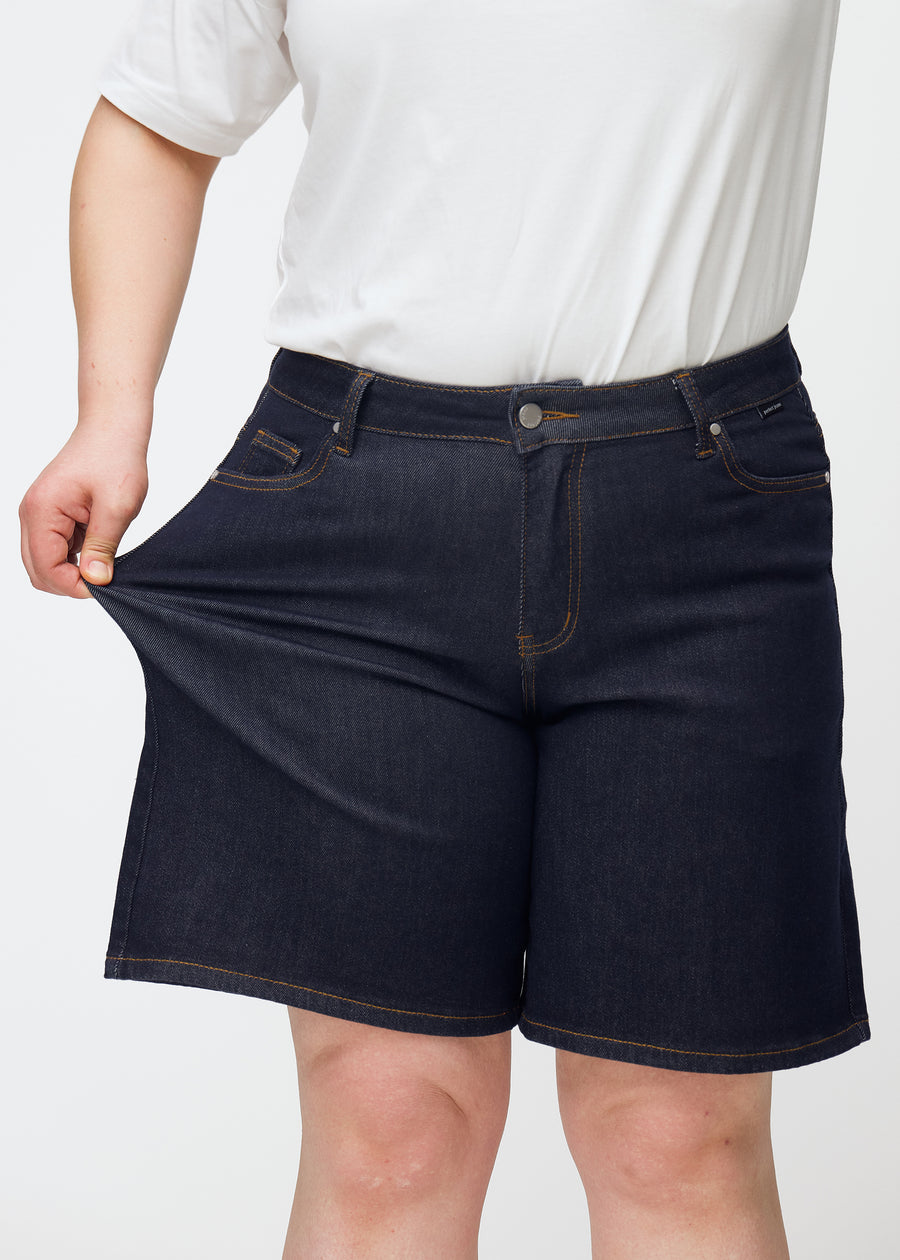 Plus-size modell strekker dyp mørkeblå denimshorts til dame i fargen Midnights ved låret for å vise stretch.