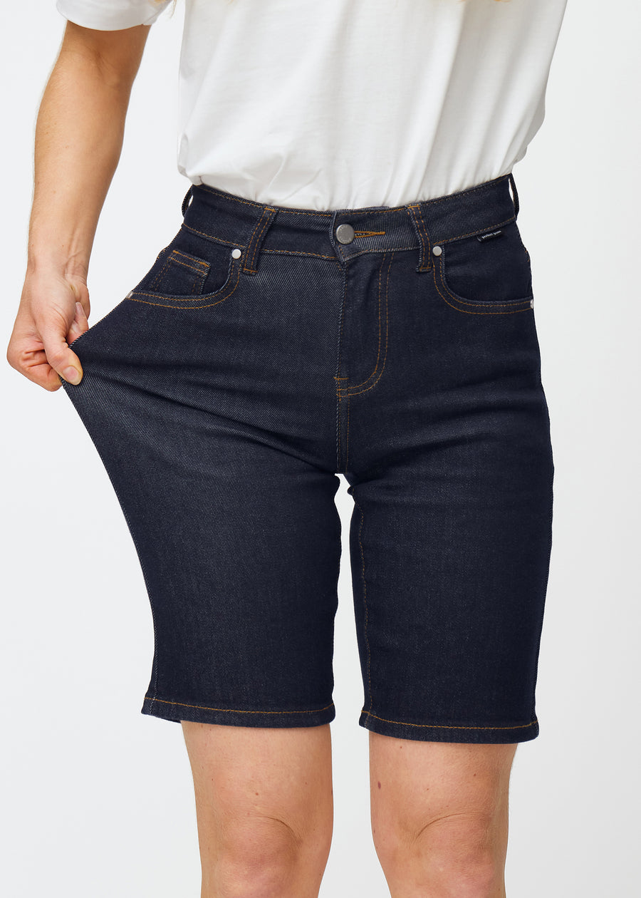 Modell strekker dyp mørkeblå denimshorts til dame i fargen Midnights ved låret for å vise materialets stretch.