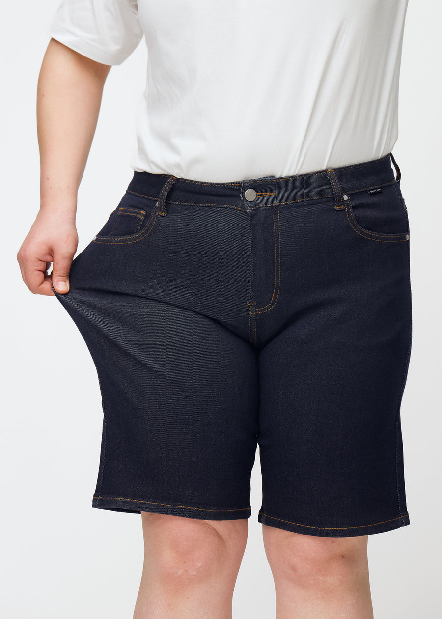 Plus-size modell strekker dyp mørkeblå denimshorts til dame i fargen Midnights ved låret for å vise stretch.