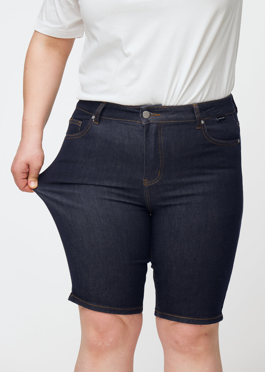 Plus-size modell strekker dyp mørkeblå denimshorts til dame i fargen Midnights ved låret for å vise stretch.