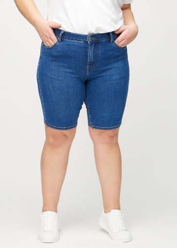 Mørk denim skinny middle shorts, modellnavn Oceans, som sitter tett på låret og slutter rett over kneet på en plus-size modell, sett forfra.