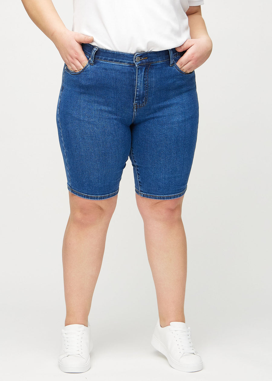 Mørk denim skinny middle shorts, modellnavn Oceans, som sitter tett på låret og slutter rett over kneet på en plus-size modell, sett forfra.