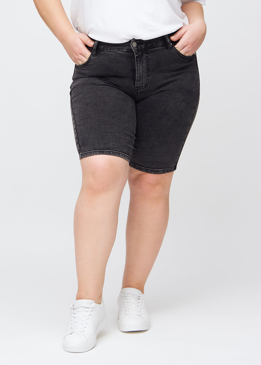Mørkegrå denim skinny middle shorts, modellnavn Shadows, som sitter tett til låret og slutter rett over kneet på en plus-size modell, sett forfra.