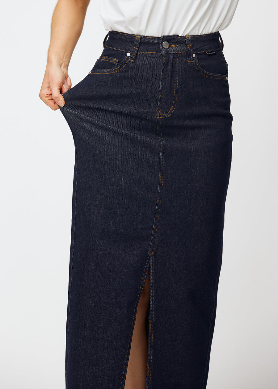 Modell strekker det lange dyp mørkeblå denimskjørtet til dame i fargen Midnights ved hoften for å vise materialets stretch.