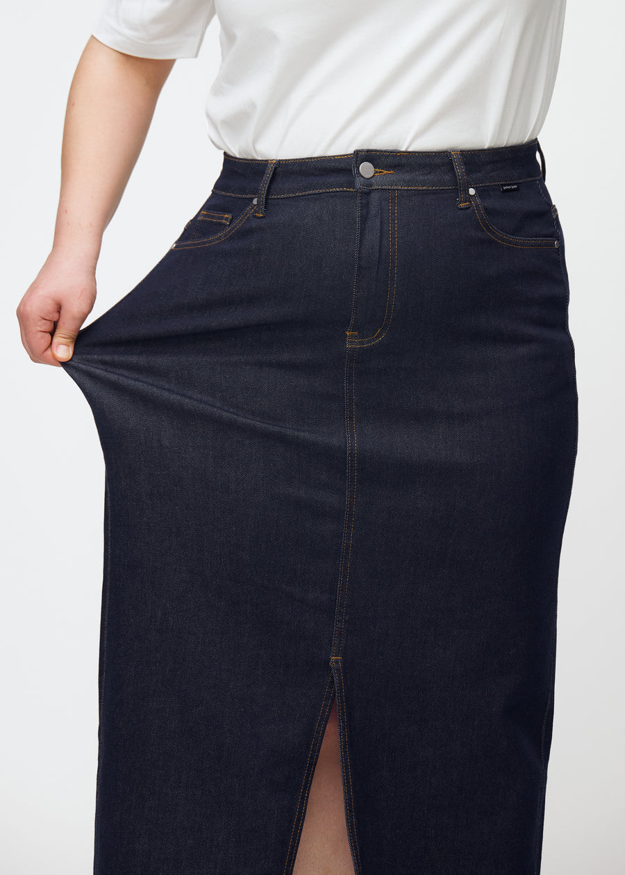 Plus-size modell strekker det lange dyp mørkeblå denimskjørtet til dame i fargen Midnights ved hoften for å vise stretch.