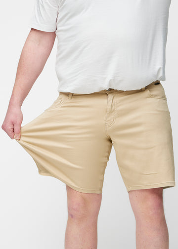 Plus-size modell strekker beige shorts til herre i fargen Gazelles ved låret for å vise materialets stretch.