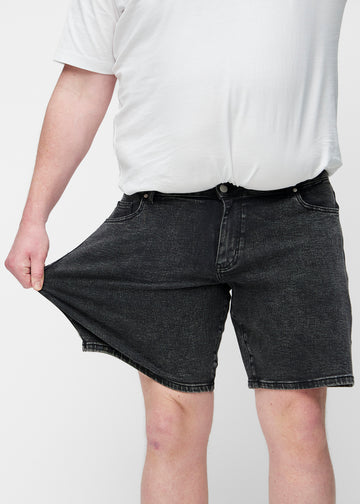 Plus-size modell strekker mørkegrå denimshorts til herre i fargen Shadows ved låret for å vise materialets stretch.