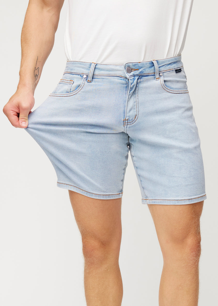 Modellen strekker lyseblå denimshorts til menn i fargen Waves ved låret for å vise materialets stretch.