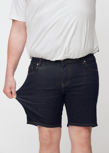 Plus-size modell strekker dyp mørkeblå denimshorts til herre i fargen Midnights ved låret for å vise stretch.