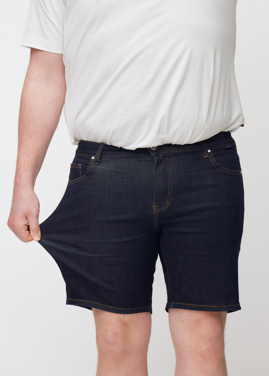 Plus-size modell strekker dyp mørkeblå denimshorts til herre i fargen Midnights ved låret for å vise stretch.